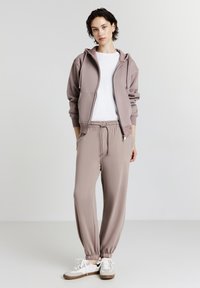 Hellbrauner Zip-Hoodie mit Kordelzug und Fronttaschen, kombiniert mit passenden Jogginghosen mit elastischen Bündchen. Darunter ein weißes T-Shirt.