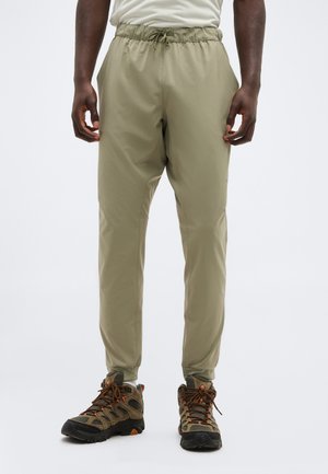 Άνδρας που φοράει μπεζ παντελόνι με ελαστική μέση και κορδόνι και καφέ πεζοπορικά παπούτσια, στέκεται μπροστά σε απλό ανοιχτό φόντο.