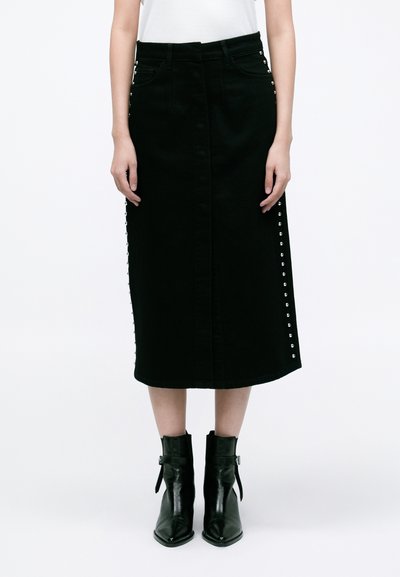 Jupe midi en denim noir avec une fermeture à boutons sur le devant, ornée de rivets argentés le long des coutures latérales. Portée avec des bottines noires.