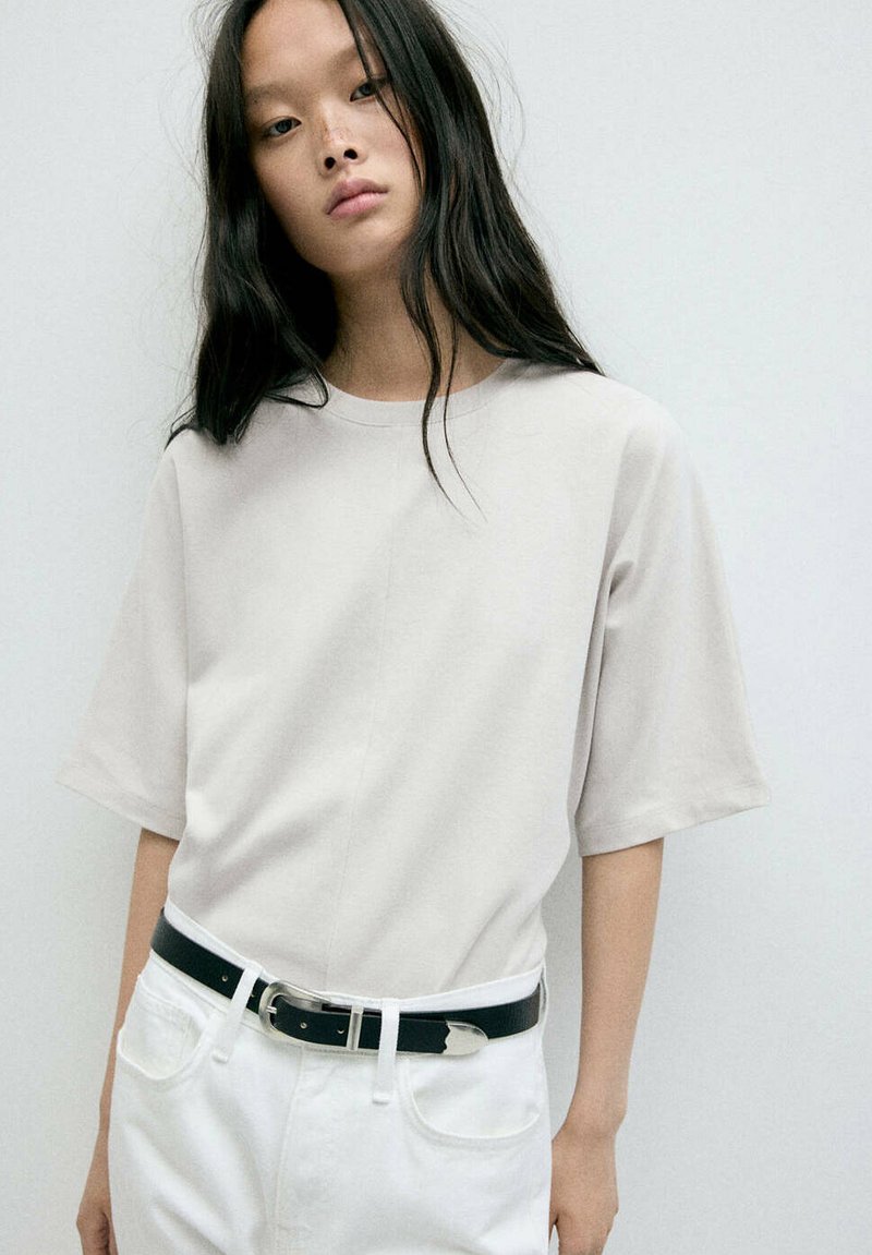Jeune femme aux longs cheveux noirs porte un t-shirt uni beige clair à manches courtes, rentré dans un pantalon blanc avec une ceinture noire, sur un fond clair.
