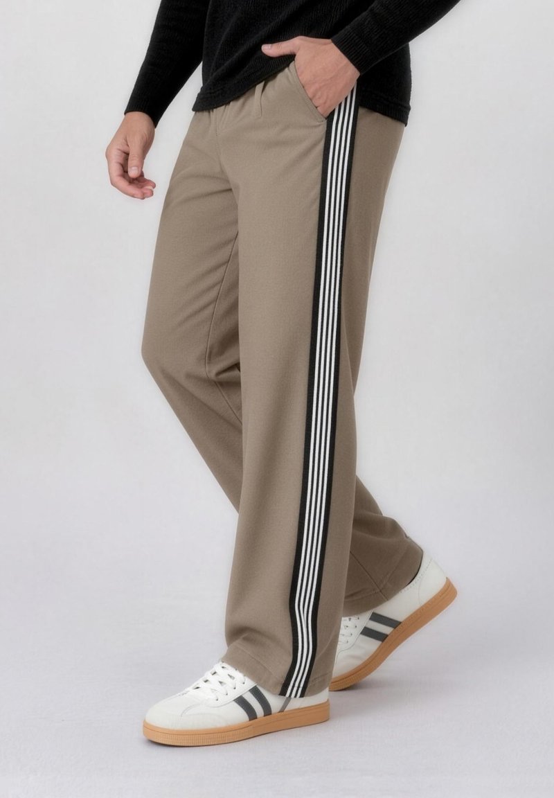 Άτομο που φοράει μπεζ παντελόνι με μαύρες και λευκές ρίγες στα πλάγια, λευκά αθλητικά παπούτσια με μαύρες ρίγες, στέκεται μπροστά σε ένα απλό φόντο.