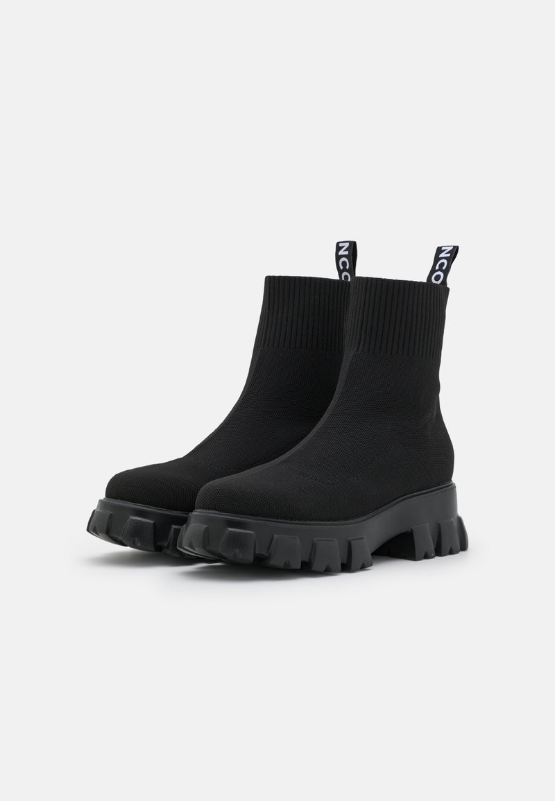 Bottines noires en tissu tricoté avec un revers à côtes, une semelle épaisse texturée et une languette portant une marque blanche.