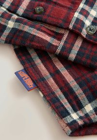 Czerwono, granatowo i biało w kratkę materiał z detalami guzików oraz fioletową metką z napisem "Superdry" w pomarańczowym kolorze.