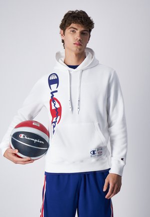 Sudadera blanca con un bolsillo delantero, que presenta un logo gráfico en rojo y azul. El modelo sostiene una pelota de baloncesto con acentos en rojo y gris.
