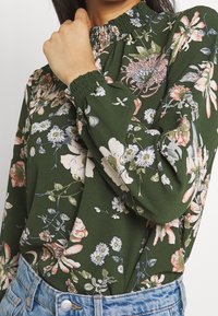 Grön blus med blommönster och smockad hög krage, prydd med rosa, vita och blå blommor; texturerad tyg och långa, samlade ärmar.