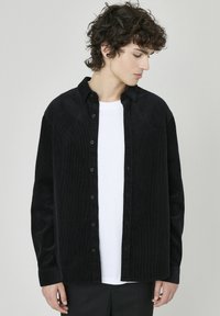 Chemise en velours côtelé noir à boutons sur le devant, avec des manches longues et un col classique, associée à un t-shirt blanc à col rond.