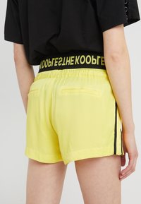 Pantaloni corti gialli con una cintura elastica nera che presenta il testo "KOOGE" e due tasche posteriori. Tessuto liscio e leggero con strisce laterali.