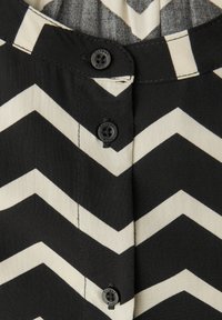 La tela a rayas en zigzag negra y crema cuenta con seis botones negros a lo largo del placket frontal, con detalles de costura visibles.