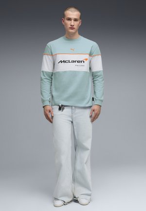 Jeune homme portant un sweat-shirt McLaren Racing turquoise et blanc, un jean large bleu clair et des baskets blanches, debout devant un fond gris.