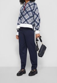 Felpa oversize a quadretti blu marino con orlo bianco e cordino rosso, abbinata a pantaloni blu marino dalla vestibilità comoda e mocassini neri, mentre tiene una borsa blu marino.
