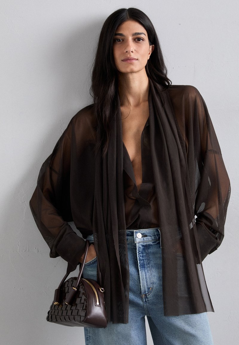 Top nero trasparente con scollatura profonda a V e maniche lunghe, abbinato a jeans chiari strappati e una borsa intrecciata scura con cerniera.