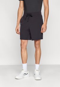 Črne kopalne kratke hlače z elastiko za vezavo v pasu, stranskimi žepi in gladkim, lahkim materialom. Nošene z belimi športnimi čevlji in nogavicami.