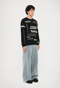 Čierny sveter s logom "ICECREAM" v rôznych písmach, doplnený širokými svetlomodrými kordovými nohavicami. Outfit dopĺňajú čierne topánky.