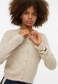 Béžový žebrovaný cardigan s kulatým výstřihem, zdobený knoflíky na přední straně a dlouhými rukávy. Na zápěstí stříbrný náramek.