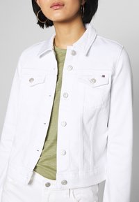 Nainen, jolla on päällä valkoinen farkkutakki, jossa on hopeiset napit, kultaiset rengaskorvakorut ja oliivinvihreä paita sen alla.