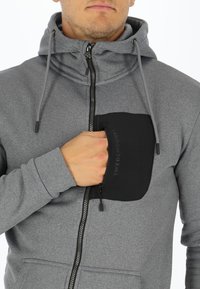 Grijze hoodie met een ritssluiting aan de voorkant, een zwart patchzakje, verstelbare koordjes en een zachte textuur. Logo zichtbaar op de zak.