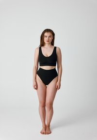 Frau steht barfuß in schwarzer, hoch taillierter Unterwäsche und passendem Crop-Top vor einem schlichten weißen Hintergrund.