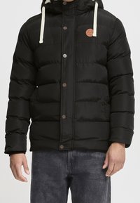 Mann trägt eine schwarze gesteppte Winterjacke mit Knöpfen, zwei Taschen vorne, einer Kapuze und weißen Kordeln, kombiniert mit dunklen Jeans.
