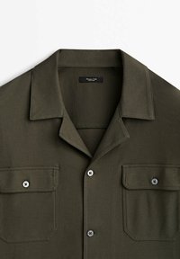 Chemise boutonnée vert foncé avec deux poches poitrine à rabat et un col large, en tissu texturé.