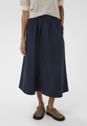 Femme portant une jupe midi bleu marine avec une ceinture élastique et des sandales en daim marron avec des semelles noires.