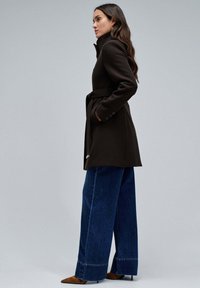 Abrigo de mezcla de lana marrón con cuello alto, cintura ajustada con cinturón y bolsillos laterales, combinado con jeans de pierna ancha y tacones marrones de punta.