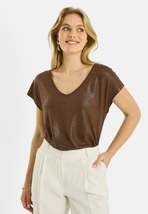 Mujer sonriente con una blusa marrón de manga corta y escote en V y pantalones blancos de talle alto, con una mano en el bolsillo, sobre un fondo blanco.