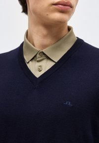 Άνδρας που φοράει μπλε σκούρο πουλόβερ με V λαιμόκοψη πάνω από μπεζ πόλο πουκάμισο με κουμπιά, με εμφανές το κάτω μέρος του προσώπου και το άνω μέρος του στήθους.