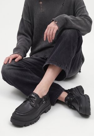 Person, der sidder med benene i kors, iført sorte chunky platformssko, løse sorte bukser og en mørkegrå slidt sweater.