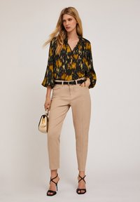 Blusa a maniche lunghe con un motivo ikat giallo e nero, abbinata a pantaloni beige a sigaretta e tacchi neri con cinturini, mentre tiene una borsa metallica.