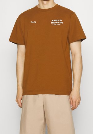 Uomo che indossa una maglietta marrone con il testo "foret" e "A WALK IN THE WOODS" abbinata a pantaloncini beige su uno sfondo semplice.