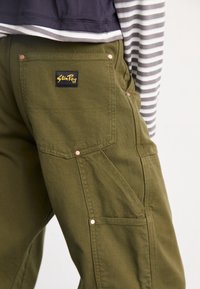 Pantalon vert olive avec plusieurs poches cousues et rivets en cuivre, avec une étiquette noire et jaune "Stan Ray" sur la poche arrière.