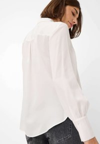 Blusa bianca con maniche lunghe a sbuffo e colletto classico. Presenta una leggera lucentezza e un orlo curvo. Il dettaglio della piega sul retro aggiunge dimensione.