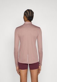 Garām piedurknēm sporta krekls mīkstā rozā audumā ar augstu apkakli, aizmugures šuves detaļu un slaidu piegriezumu. Kombinēts ar tumši sarkaniem šortiem.