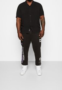 Zwart shirt met korte mouwen en knoopsluiting; gecombineerd met zwarte sweatpants met witte en paarse graphics. Witte sneakers maken de look compleet.