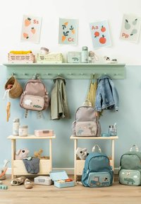 Sacs à dos et vestes d’enfants suspendus à des crochets au-dessus d’étagères en bois avec des jouets et des boîtes à lunch sous des affiches de l’alphabet avec des fruits et légumes.