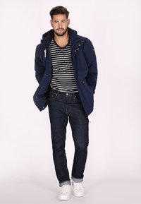 Giacca a vento blu navy con cappuccio sopra una maglietta a righe bianco e nera, abbinata a jeans in denim scuro e sneaker bianche.