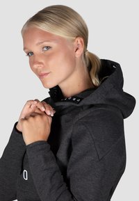 Charcoal-Hoodie mit hohem Kragen, verkörpert ein Logo auf der Vorderseite, weiche Textur und enge Passform, zeigt flache Nähte und eine Kordel.