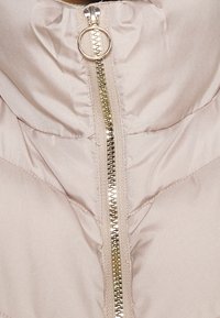 Beige prošívaná bunda se zlatým zipem a kulatým úchytem, detail na límec a oblast hrudi.