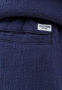 Tumši zila teksturēta auduma ar krinkļotu rakstu; tam ir balta birka ar uzrakstu "JACK & JONES EST. 1990", kas ir iestrādāta.