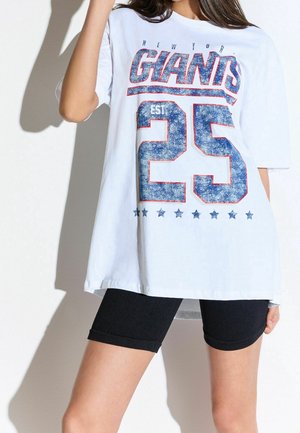 Frau trägt ein übergroßes weißes T-Shirt mit dem Aufdruck "New York Giants EST 25" in Blau und Rot, kombiniert mit schwarzen Shorts, vor schlichtem Hintergrund.
