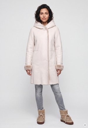 Frau trägt einen hellbeigen Kapuzenmantel mit Fellmanschetten, graue Jeans und beige Winterstiefel und steht vor einem schlichten hellen Hintergrund.