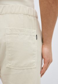 Κοντινό πλάνο της τσέπης μπεζ παντελονιού με μικρή μαύρη ετικέτα στη δεξιά πλευρά, δίπλα στο χέρι και το μπράτσο ενός ατόμου.