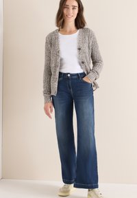 Cárdigan gris de punto con dos botones, camiseta blanca de cuello redondo y jeans oscuros de pierna ancha. Zapatillas crema completan el conjunto.