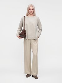 Femme aux longs cheveux blonds portant un pull beige et un pantalon beige, tenant un sac à bandoulière en cuir marron, debout devant un fond blanc.