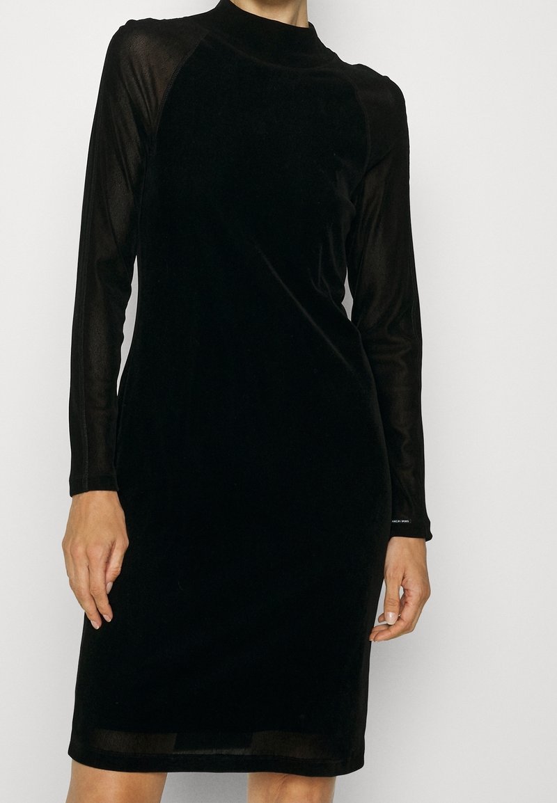 Femme portant une robe en velours noir ajustée avec de longues manches transparentes et un col montant, debout devant un fond blanc uni.
