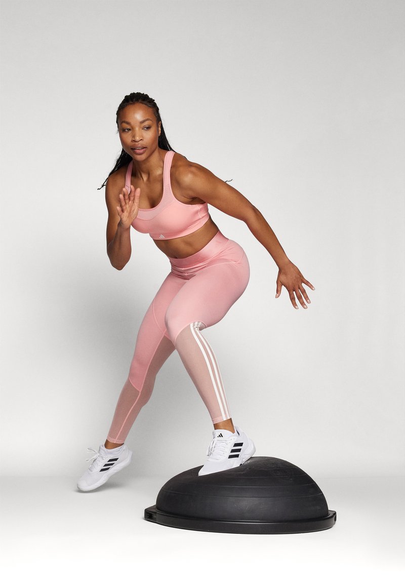 Soutien-gorge de sport rose et leggings avec des rayures blanches, associés à des chaussures de sport blanches, en train de faire un exercice sur un entraîneur d'équilibre noir.