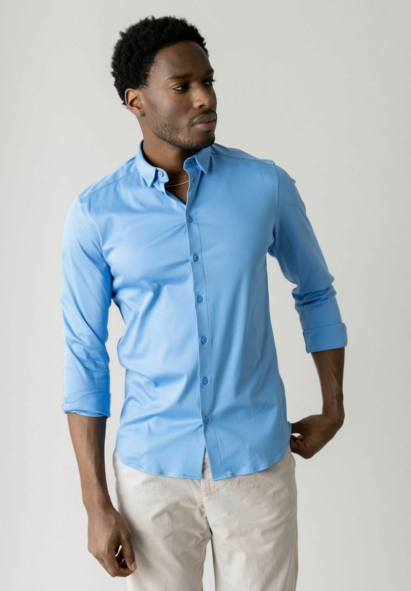 Vīrietis regulē gaismas zilas pogām aizdares kreklu ar uzruļļotām piedurknēm, valkājot gaišu bikses uz vienkārša fona.