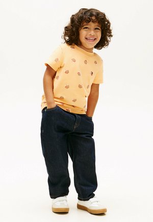 Uśmiechnięte dziecko o kręconych włosach stoi z rękami w kieszeniach, ubrane w pomarańczową koszulkę z motywem koszykówki, ciemne dżinsy i białe trampki na białym tle.