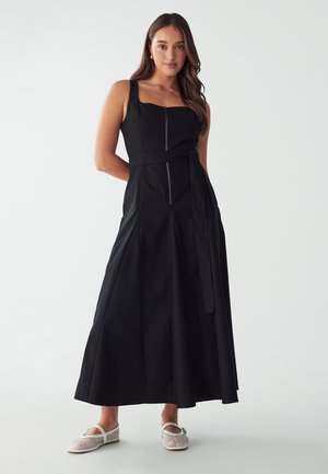 Black, sleeveless maxi dress with square neckline, front zipper, waist tie, and pleated skirt. Model is wearing white mesh flats.