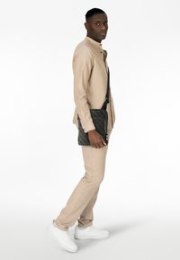 Borsa messenger nera con texture e design a motivo, indossata a tracolla, abbinata a una camicia di lino beige e pantaloni coordinati. Scarpe da ginnastica bianche.